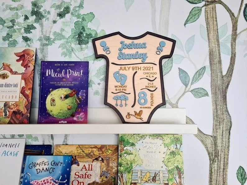 Wooderland personalized baby boy birth announcement sign in a onesie shape with blue accents on a nursery shelf.