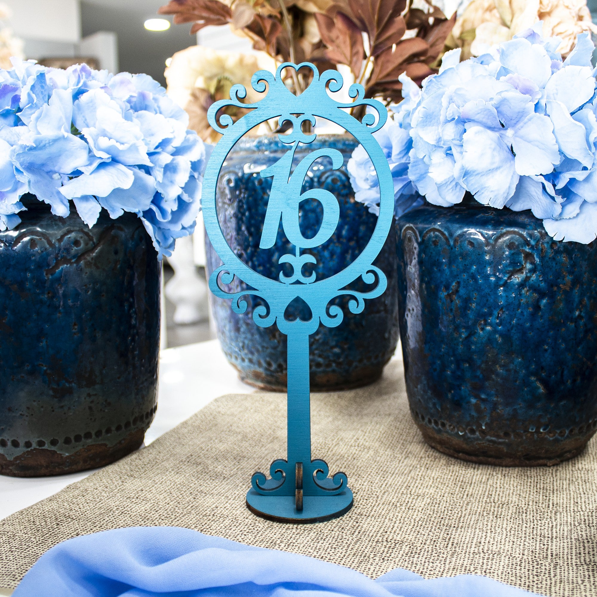Wooderland blue wooden table number 16 with ornate circular frame on a burlap table runner with blue hydrangea decor.