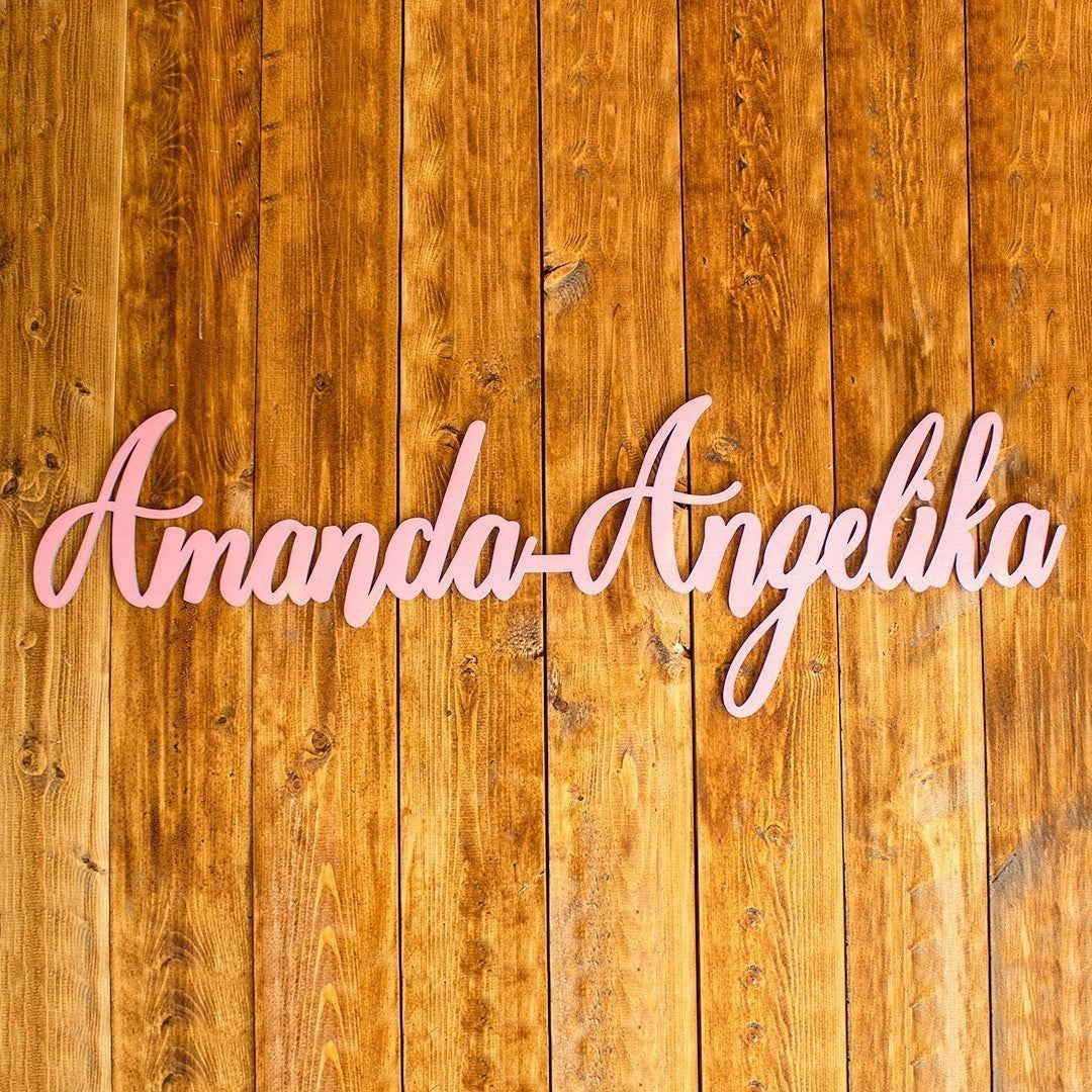 Wooderland custom laser-cut wooden name sign in blush pink script font mounted on a rustic vertical wood backdrop.