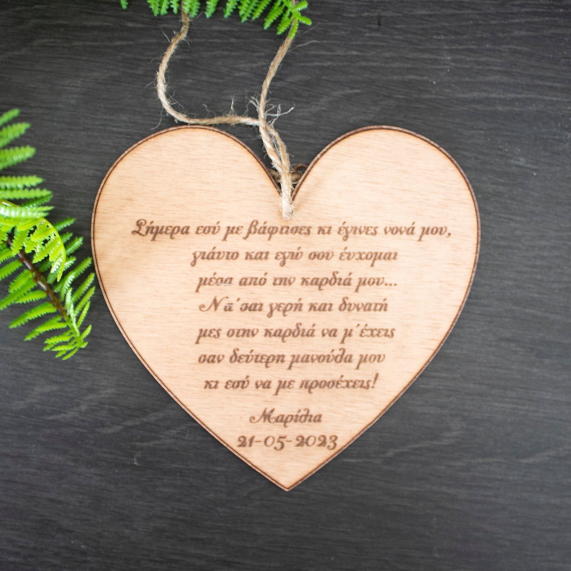 Wooderland wooden heart keepsake engraved with a Greek christening message for a godmother, hanging on a dark surface.