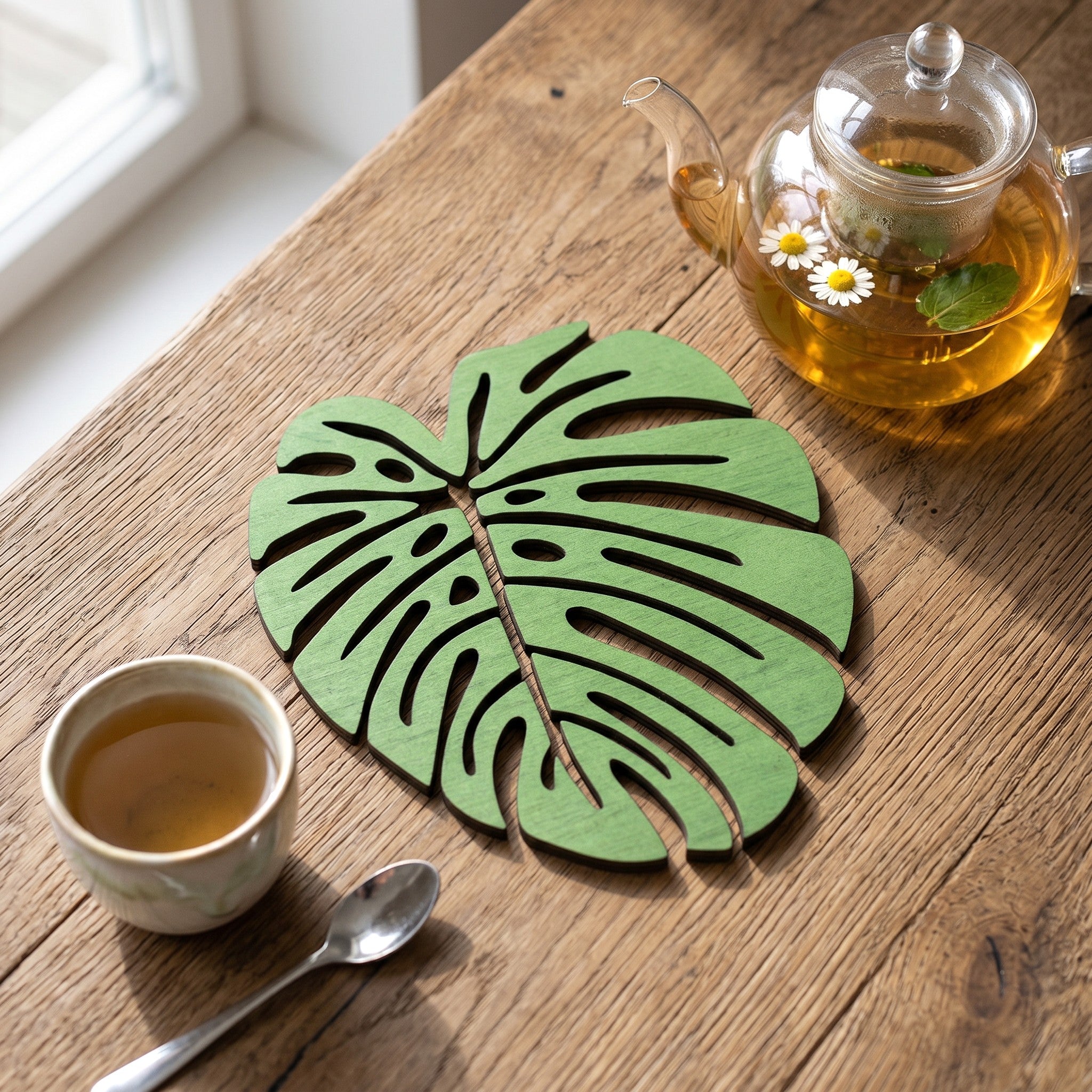 Wooderland green monstera leaf shaped plywood coaster on a rustic wooden table with a glass teapot and tea cup.