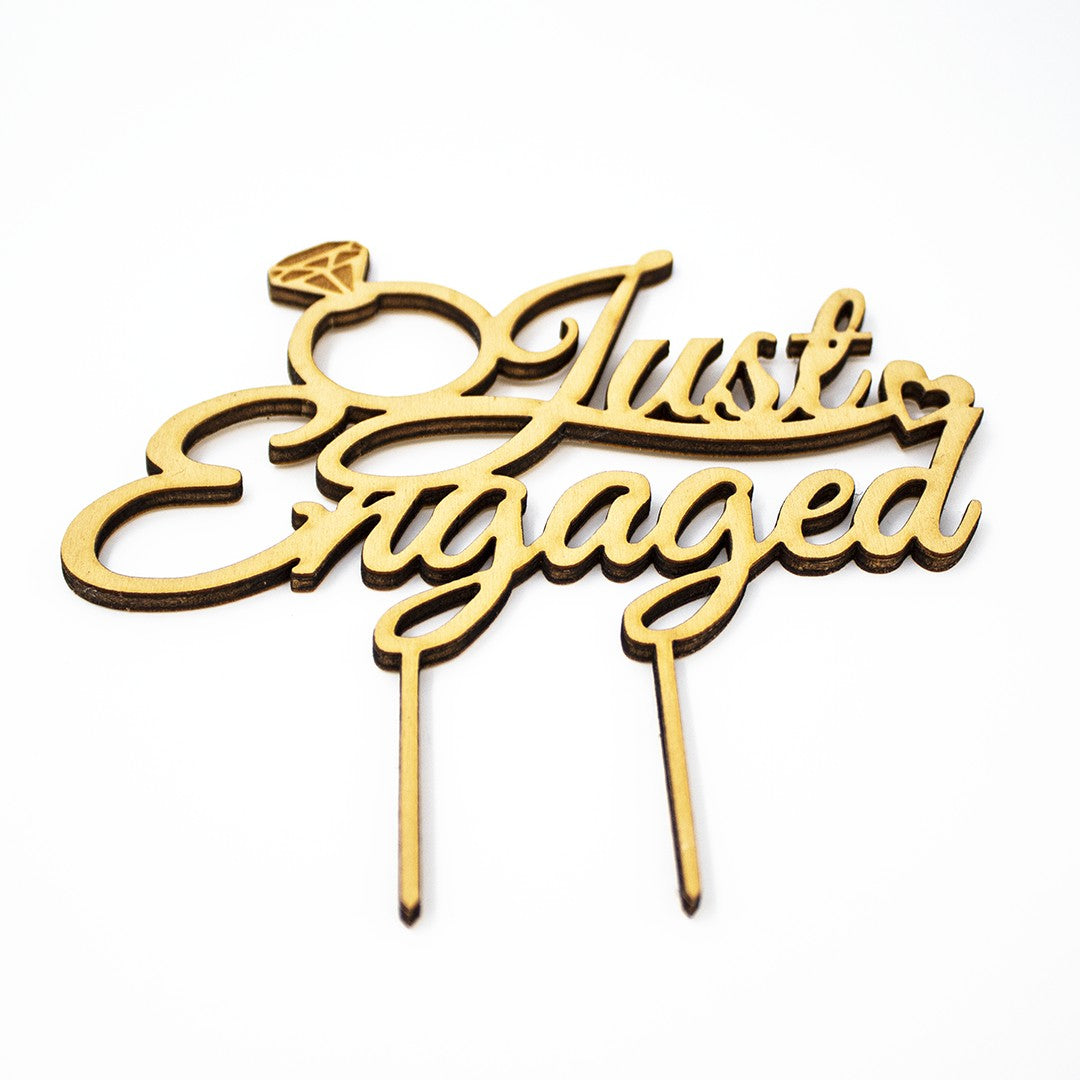 Wooderland Just Engaged wooden cake topper with laser-cut script, diamond ring, and heart detail on white background.