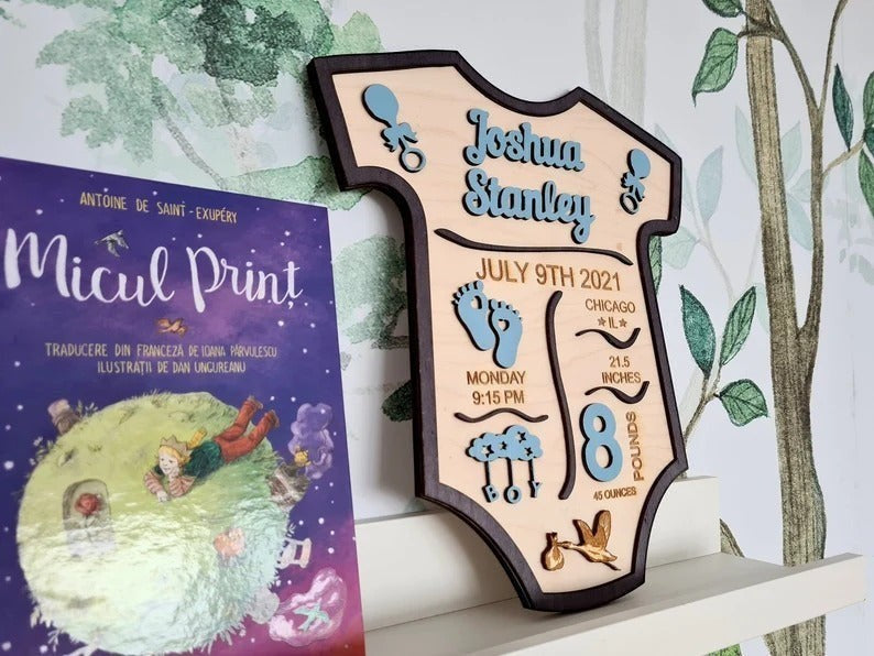 Wooderland personalized baby boy birth announcement sign shaped like a onesie with blue wooden details on a shelf.