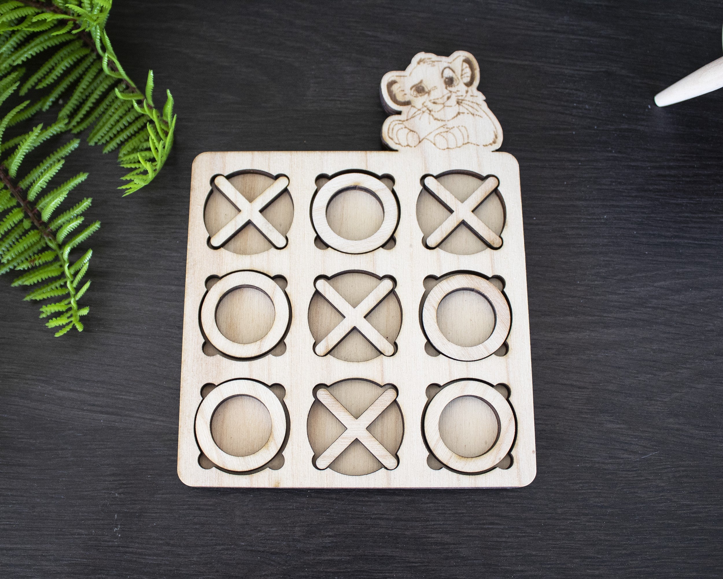 Wooderland custom wooden tic-tac-toe game board with laser-engraved lion cub figure on a dark surface.